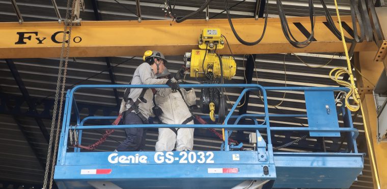 Técnicos con arnés inspeccionando el polipasto de una grúa puente desde una plataforma